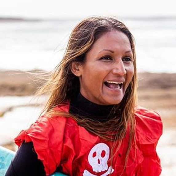 Photo of Ashika, a 41-year-old person (she/her) with dark brown hair, blonde highlights and brown eyes. Ashika is wearing a high neck black rash vest underneath a red short sleeved shirt with a white skull and cross bones logo. Ashika is pictured holding a blue surfboard on beach background.