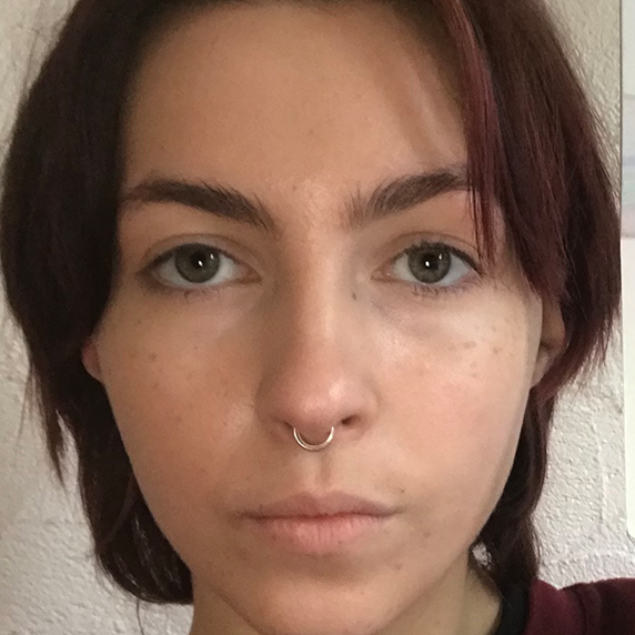 Photo of Jet, a 22-year-old person (they/them) with dark brown short hair with purple highlights and green eyes. Coco has a silver septum ring and is pictured with a light beige background.