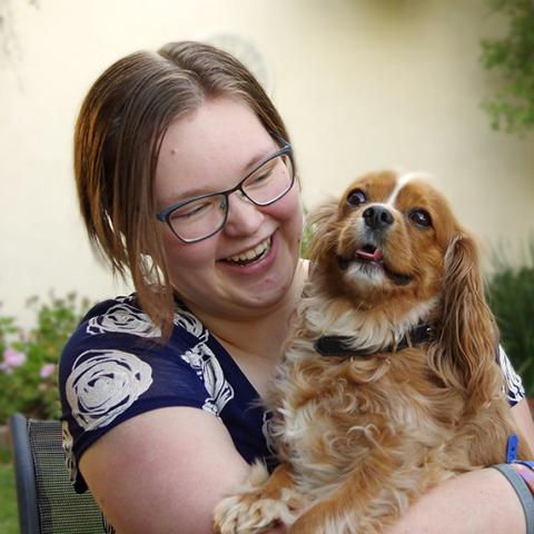 Photo of Jessi a 21-year-old person (she) with dark brown short hair with amber highlights. Jessi is smiling looking at her brown and white cocker spaniel. She is wearing blue rimmed glasses, a navy short sleeved dress with white roses on it and a collection of coloured bracelets. Jessi is pictured sitting on a chair in the garden with green and pink plants in the background.