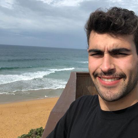 Photo of Josh, a 20-year-old person (he/him) with short black hair. Josh is wearing a black t-shirt, and is standing at a wooden lookout with a beach in the background.