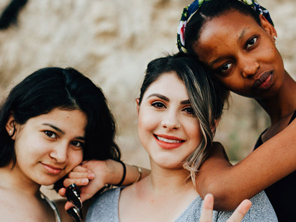 Three young girls of diverse backgrounds looking at the camera smiling, one of them doing the peace sign