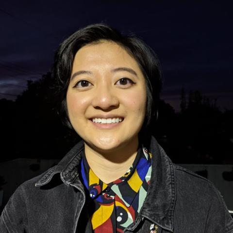 Photo of Danica, a 21-year-old old person (she/her) with dark brown short hair and brown eyes. She is wearing a black denim jacket over a collared shirt with a blue, mustard, lime and brown abstract print. Danica is pictures smiling outdoors at night with trees in the background. 