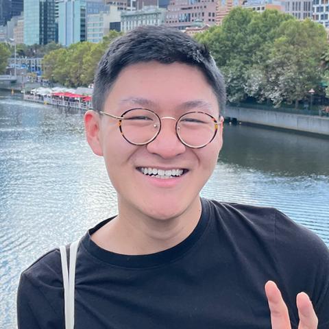 Photo of Mark, a 22-year-old person (he/him) with short black and grey hair. Mark is smiling at the camera, wearing round glasses and holding his hand up in a peace symbol. Mark is photographed on the Flinders Street bridge with the Yarra River in his background.