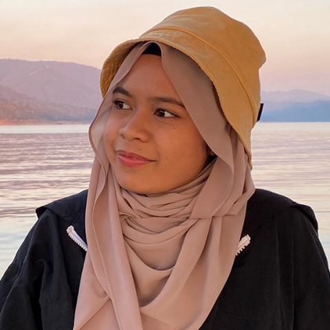 Photo of Zahra, a 20-year-old person (she/her) wearing a light pink hijab and mustard yellow bucket hat. Zahra is wearing a black weatherproof jacket with white zip. Zahra is pictured in front of a large lake with mountains and trees in the background.
